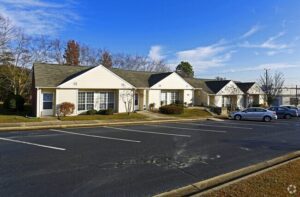 A long, single story apartment building styled to look like several attached homes with each unit alternating darker and lighter grey siding. There is a large parking lot in front