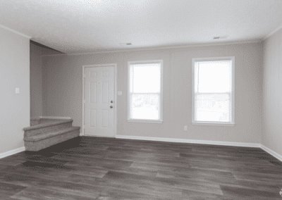 An empty living room with grey floors. The view is facing the front door and there are stairs next to the door leading to the second floor