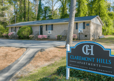 A single story triplex with a light brick exterior surrounded by trees and bushes. A sign in front declares this part of the Claremont Hills apartment community by Elevate Capital Group.