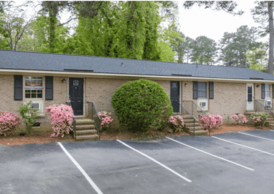 A single story triplex with a light brick exterior surrounded by trees and bushes.