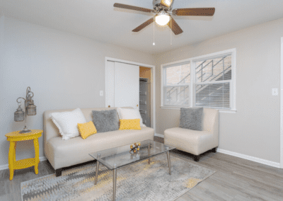 A living room staged with a creme colored couch and chair, and a glass top coffee table. There is a large closet behind the couch. We can see the entry door to the right