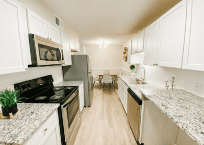 A galley-style kitchen with white cabinets, marbled white granite countertops, and stainless steel appliances. Beyond the kitchen, we see a dining table with white upholstered chairs