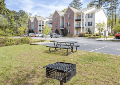 A lawn area with a park-style barbecue and a metal picnic bench in front of a large three-story apartment building at Woodland Ridge apartments