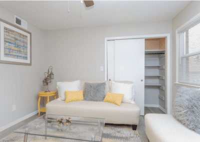 A living room staged with creme colored couches and a glass top coffee table. There is a large closet behind one couch